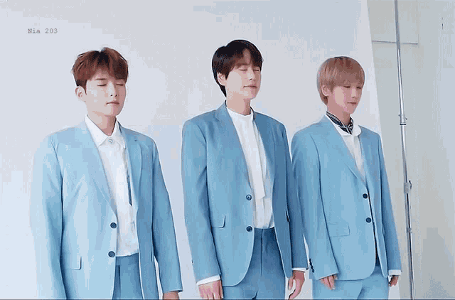 three men in blue suits are standing in front of a white wall with the number 203 on the bottom