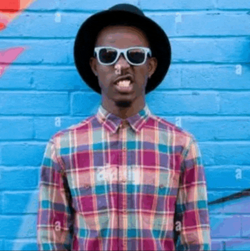 a man wearing a hat and sunglasses is sticking his tongue out in front of a colorful brick wall .
