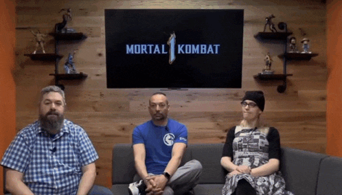 three people are sitting on a couch in front of a tv screen that says mortal kombat