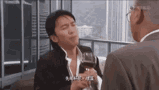 a man in a suit is holding a glass of red wine while talking to another man .