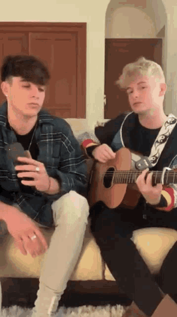 two boys are sitting on a couch playing guitars and one is holding a cell phone .