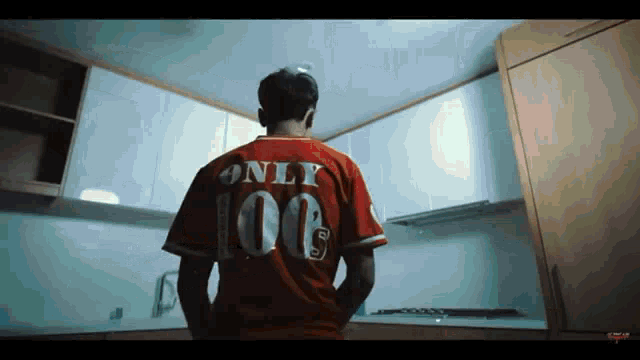 a man in a red jersey with the number 100 on the back is standing in a kitchen .