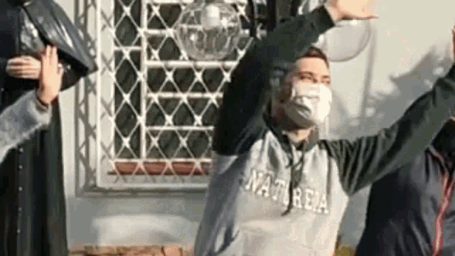 a man wearing a face mask is standing in front of a window with his arms in the air .
