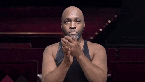 a bald man with a beard wearing a black tank top and a necklace with the letter t on it