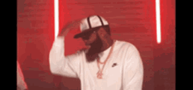 a man with a beard wearing a baseball cap and sunglasses is standing in front of a red wall .