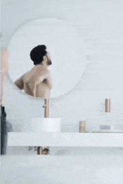 a shirtless man adjusts his belt in front of a mirror