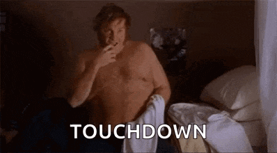 a shirtless man is sitting on a bed smoking a cigarette and taking off his pants .