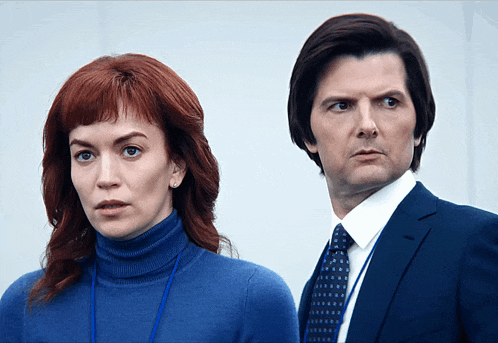 a man in a suit and tie stands next to a woman in a blue turtleneck