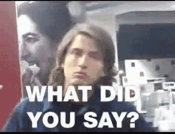 a man with long hair is standing in front of a poster that says what did you say ?