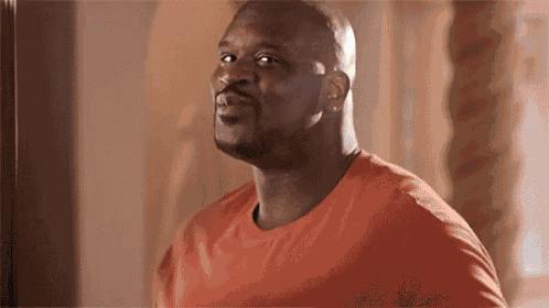 a bald man in an orange shirt is standing in a room and looking at the camera .