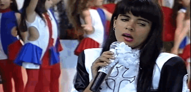 a woman is singing into a microphone while standing in front of a group of cheerleaders .