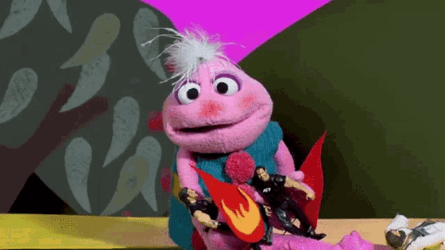 a pink puppet is playing a guitar with two action figures behind her