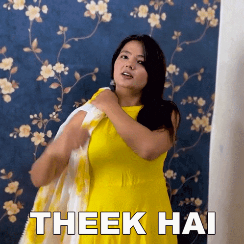 a woman in a yellow dress is standing in front of a wall with flowers on it and says theek hai