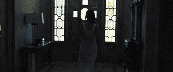 a woman in a white dress is standing in a dark room holding a door open .