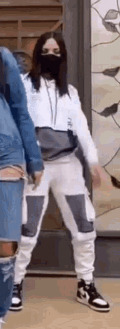 a woman wearing a mask and white pants is standing in front of a wall .