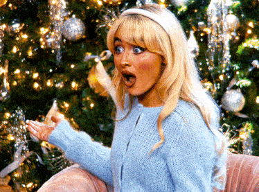 a woman in a blue sweater is sitting in front of a christmas tree with her mouth open