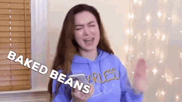 a woman in a purple hoodie is holding a bowl of baked beans in her hand .