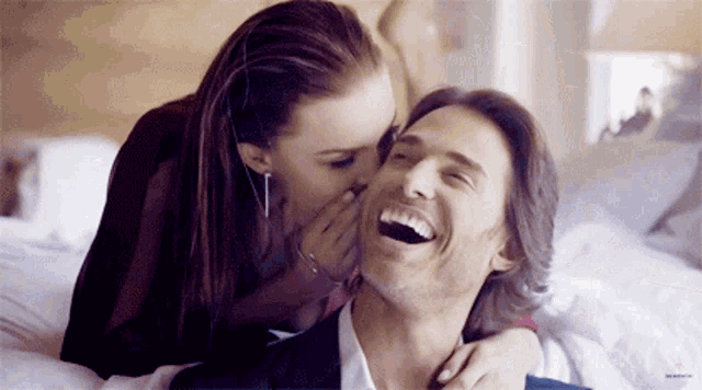 a woman whispers something into a man 's ear while laying on a bed