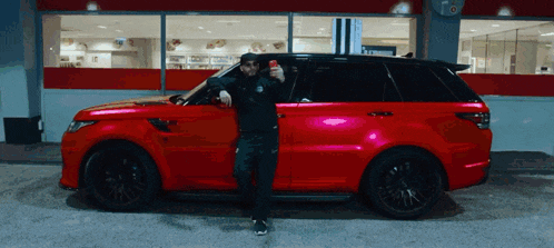 a man standing next to a red car holding a red can of soda