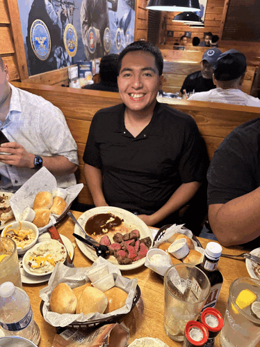 a man in a black shirt sits at a table with a plate of steak and a basket of rolls