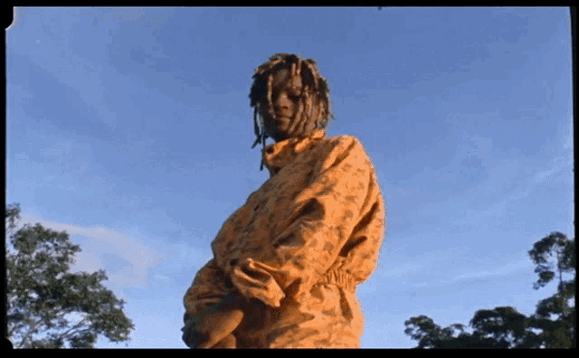 a man with dreadlocks is standing in front of trees and a blue sky
