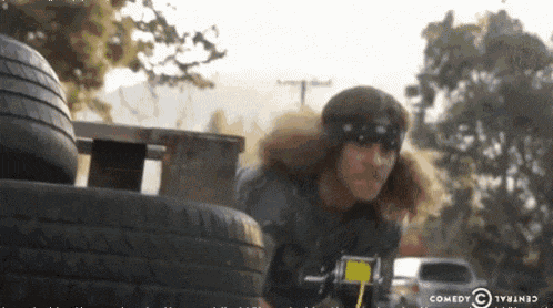 a man with long hair is standing in front of a tire and a comedy central logo