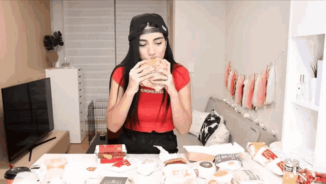 a woman wearing a red shirt that says " fucking good " is eating a hamburger