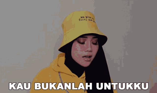 a woman wearing a yellow hoodie and a yellow hat with the words kau bukanlah untukku below her