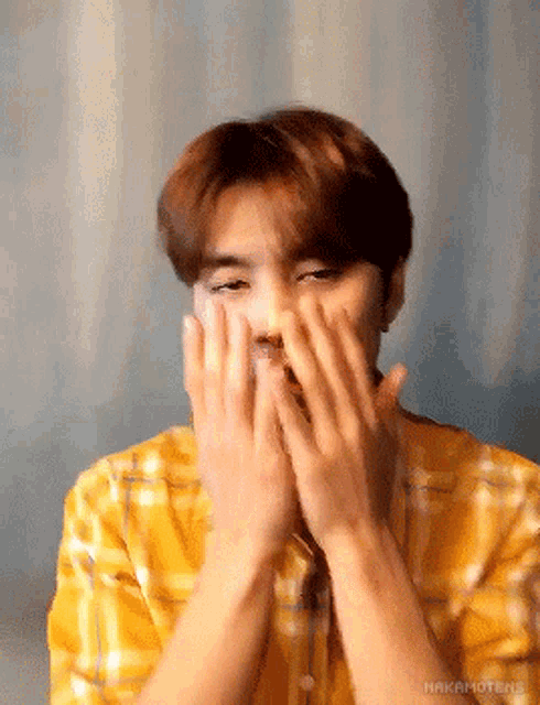 a man wearing a yellow plaid shirt is covering his mouth with his hands .