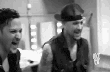 two men are standing next to each other in a room and laughing . one of the men is wearing a hat .