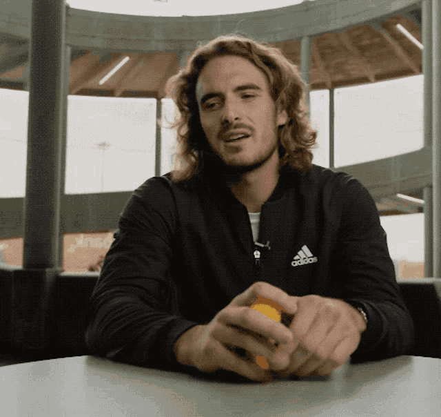 a man wearing a black adidas jacket sits at a table holding an orange