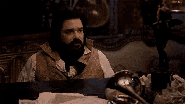 a man with a beard is sitting at a table in a dark room with a statue in the background .