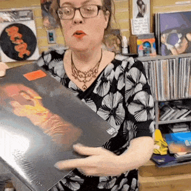 a woman wearing glasses is holding a record in front of a wall with a picture of ac dc on it