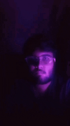 a man with glasses and a beard looks at the camera in the dark