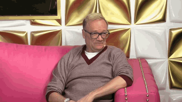a man wearing glasses sits on a pink couch in front of a gold wall