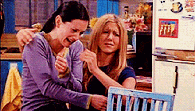 two women are sitting next to each other in a kitchen with a refrigerator in the background .