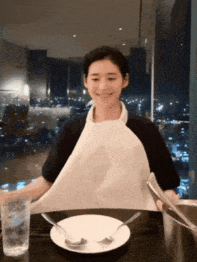 a woman wearing a white apron sits at a table with a plate and silverware