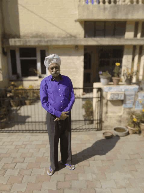 a man wearing a turban and a purple shirt stands in front of a house with the number 2976 on the side of it