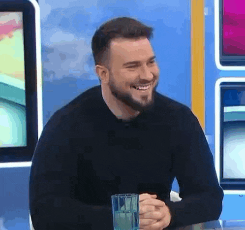 a man with a beard is smiling while sitting at a table with a glass of water in front of him .