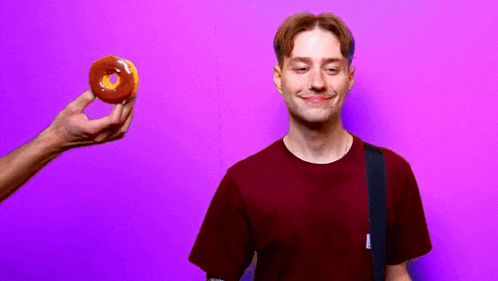 a man in a maroon shirt is smiling while a person holds a donut in front of him