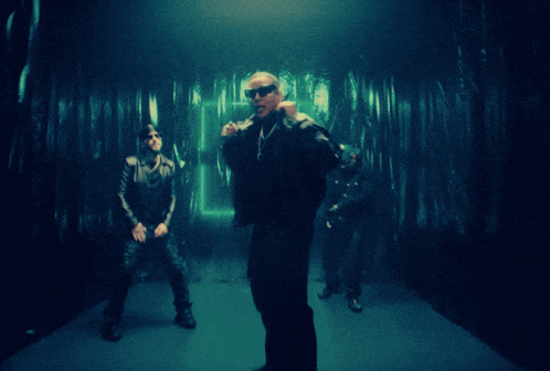 a man wearing sunglasses and a leather jacket stands in a dark room with two other men