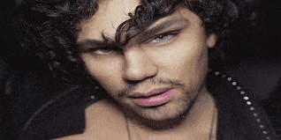 a man with curly hair and a beard is wearing a black jacket and necklace .