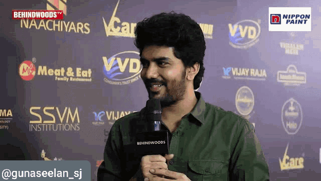 a man speaking into a microphone in front of a wall with behindwoods written on it