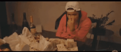 a woman in a pink hoodie is sitting at a table with a bunch of money and a bottle of wine .