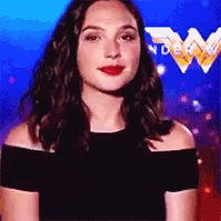 a woman wearing a black off the shoulder top is standing in front of a wonder woman logo .
