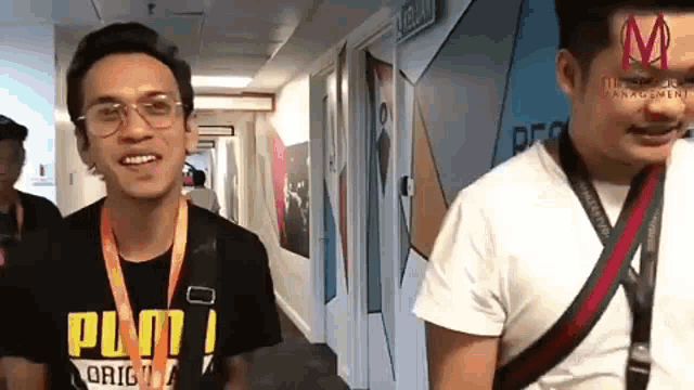 a man wearing glasses is standing in a hallway next to another man wearing a lanyard .