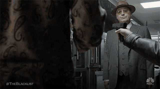 a man in a suit stands in front of a man pointing a gun at him while another man watches