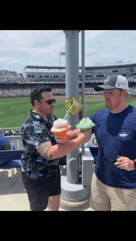 two men are standing next to each other at a baseball stadium holding ice cream drinks .