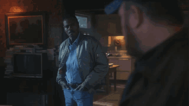 a man in a bomber jacket stands in a room with another man