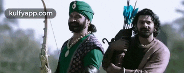 two men are standing next to each other holding bows and arrows . one of the men is wearing a green turban .